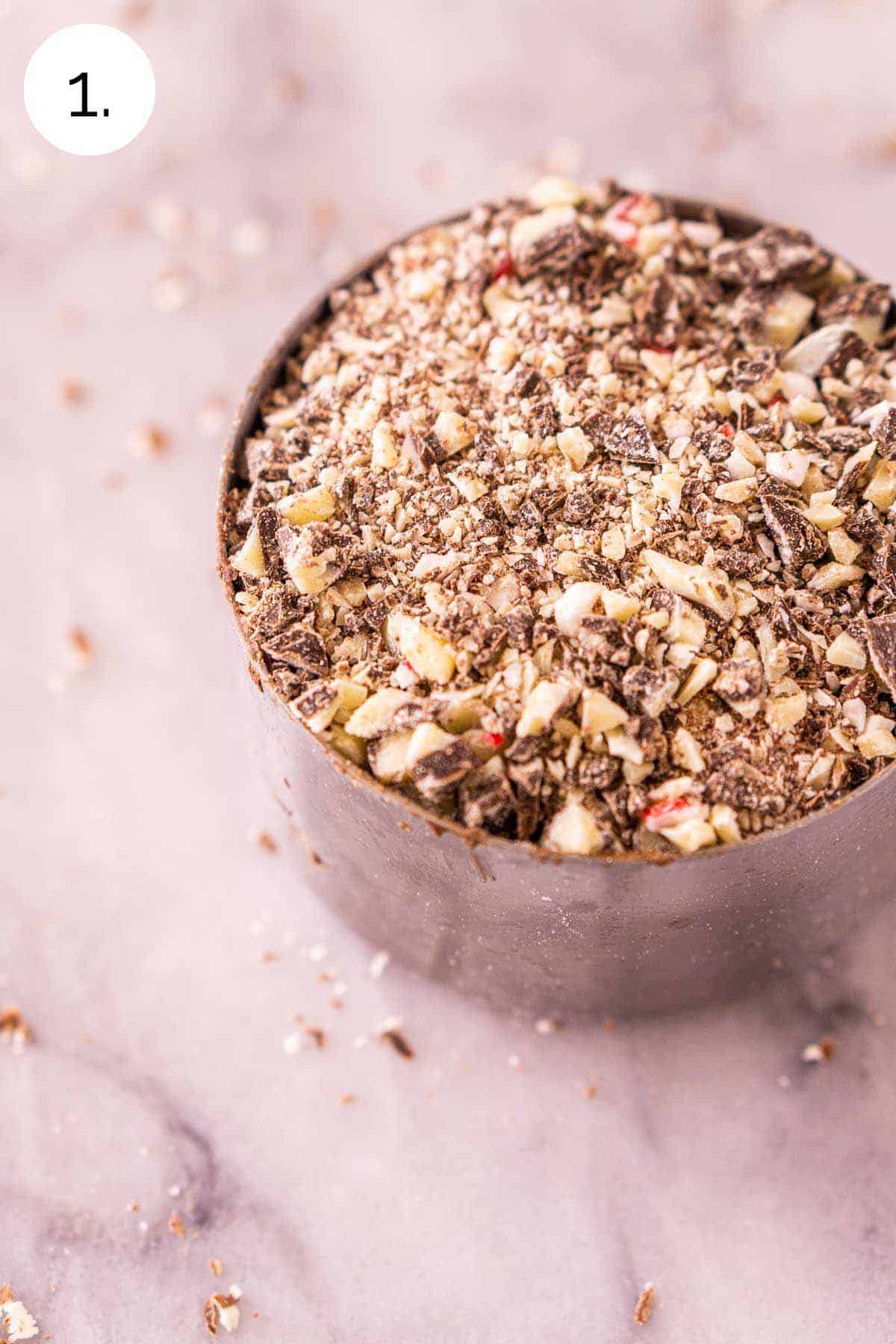 The chopped peppermint bark in a stainless steel measuring cup on a white and gray marble surface.