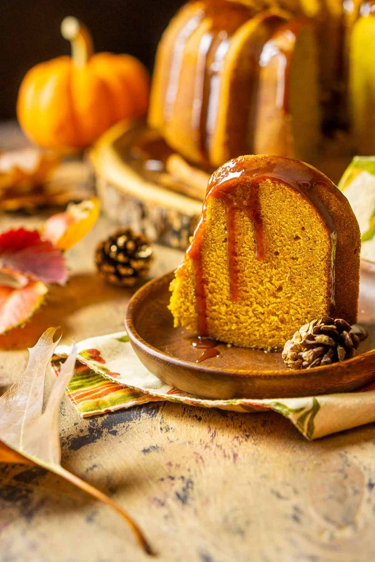 A slice of the pumpkin pound cake on a small wooden plate with the rest of the cake in the background and fall decor around it.