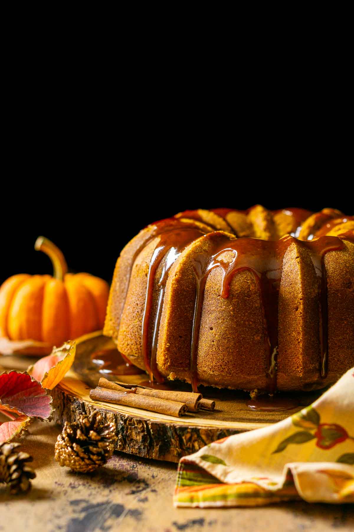 The pumpkin pound cake on a wooden serving platter against a black background with a napkin in front and fall decor surrounding it.