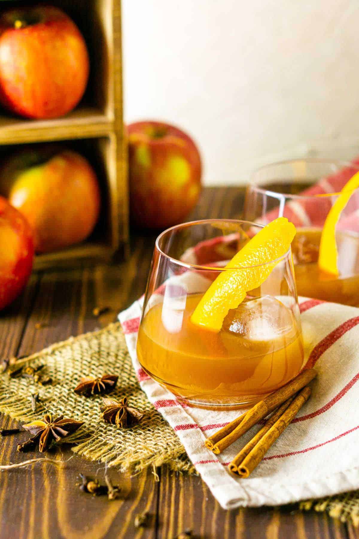 An apple cider old fashioned on a red and cream napkin with spices sprinkled around it with apples in the background.