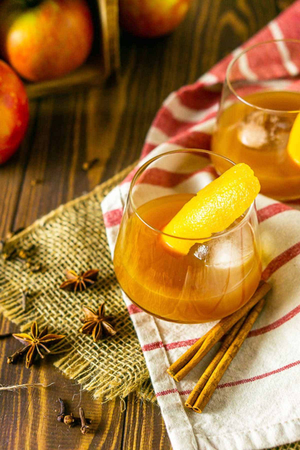 Looking down on an apple cider old fashioned on a brown wooden surface with burlap, spices and apples around it.