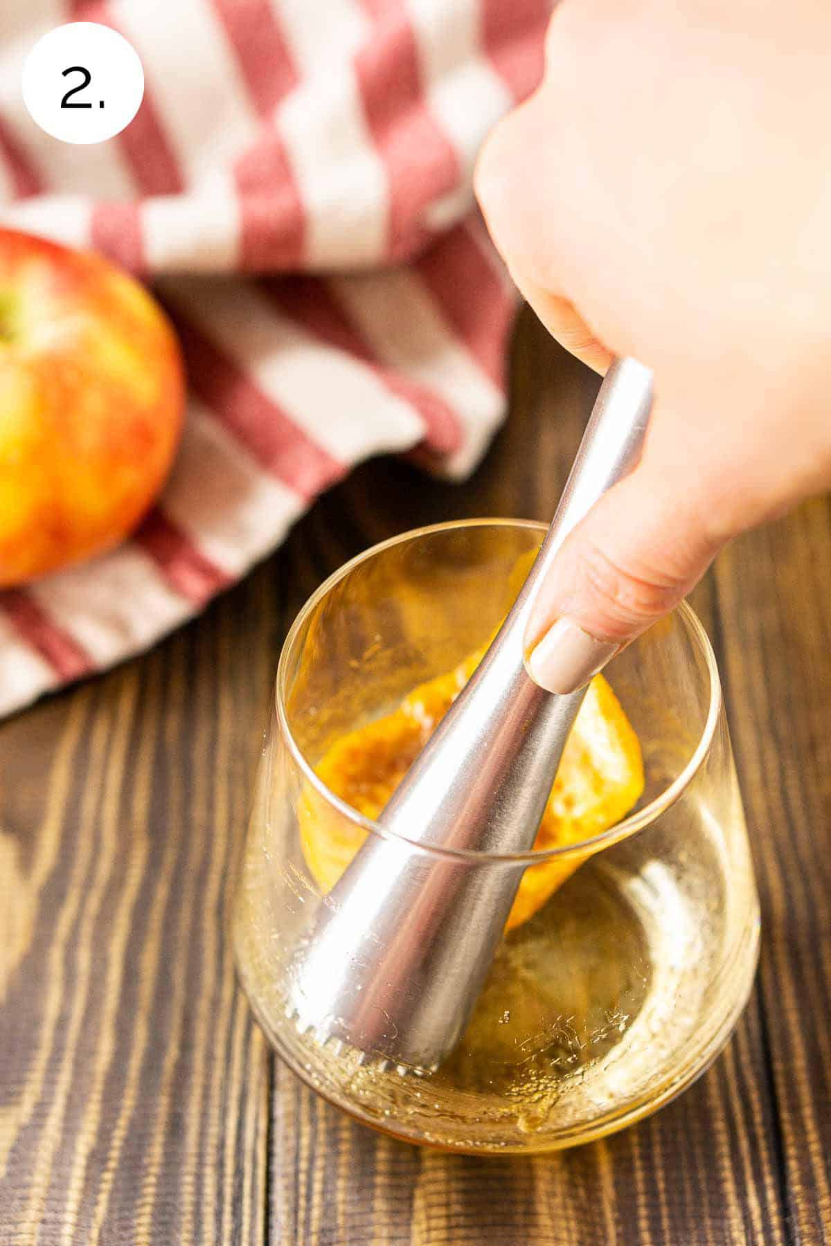 A hand holding a muddler and using it to dissolve the brown sugar in an old fashioned glass on a wooden board.