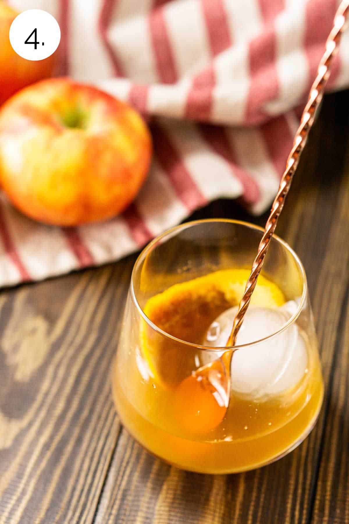 A copper bar spoon stirring the apple cider and other ingredients with a large ice cube on a brown wooden board.