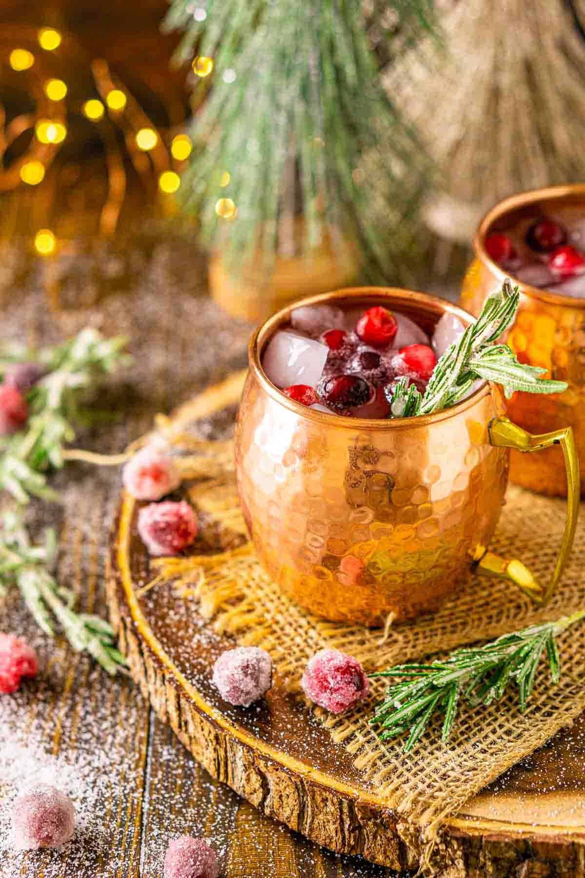 A cranberry Moscow mule on a wooden tray with holiday lights in the background and decor around it.