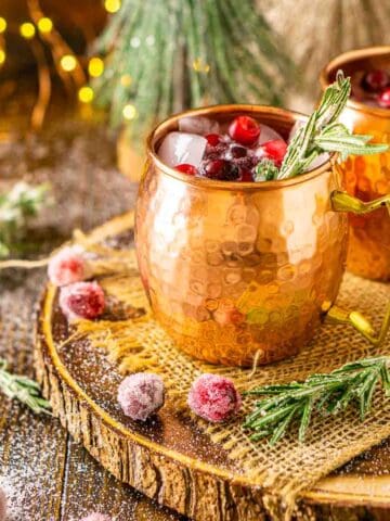 Two Christmas cranberry Moscow mule cocktails on a wooden platter with burlap and sugared cranberries and rosemary around them with lights in the background.