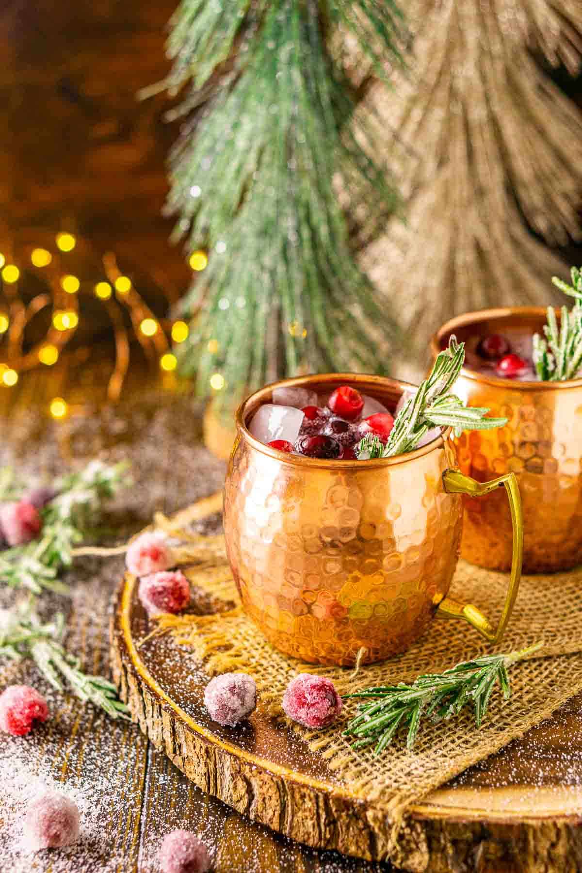 Two Christmas cranberry Moscow mule cocktails on a wooden platter with burlap and sugared cranberries and rosemary around them with lights in the background.