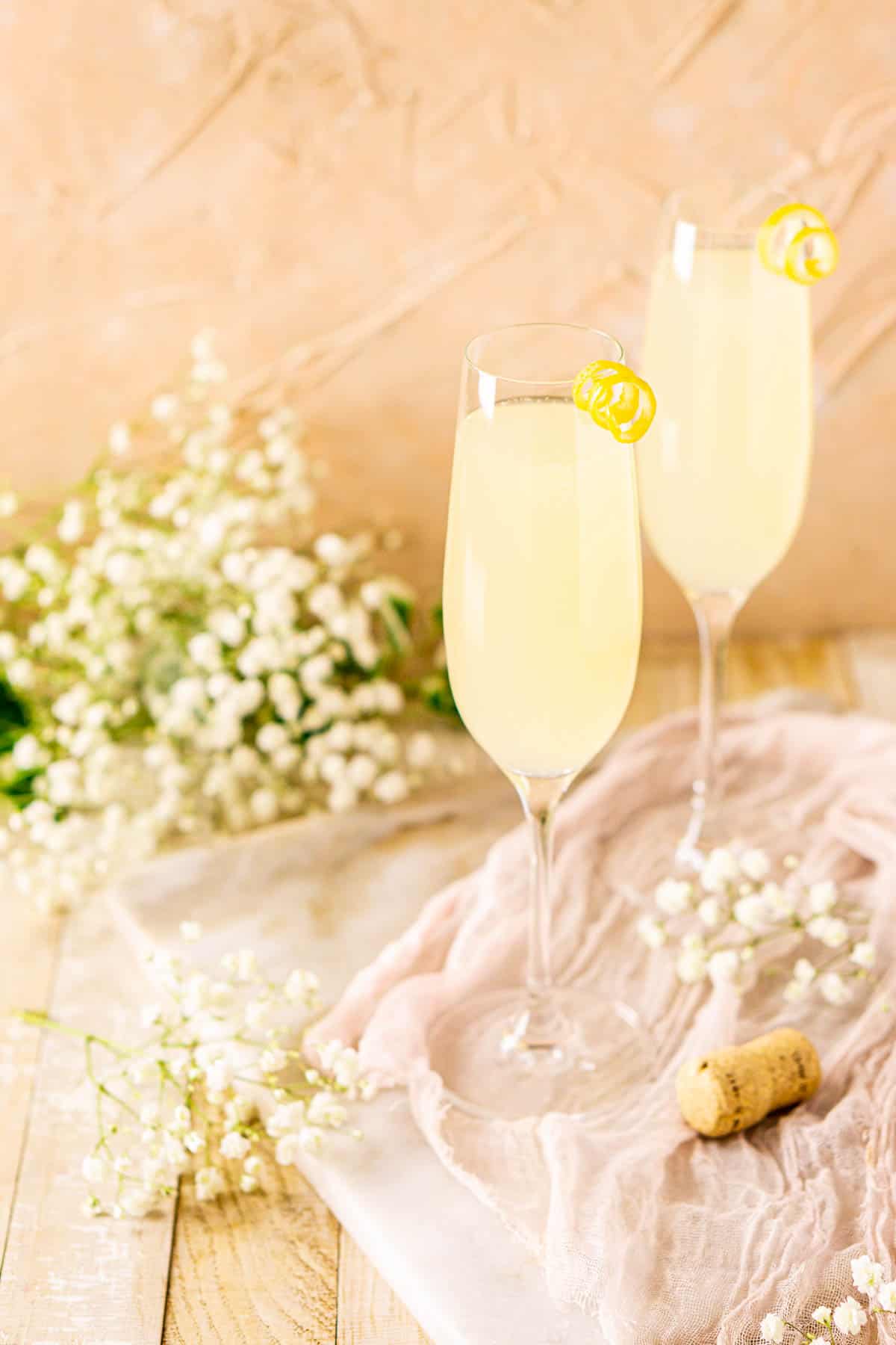 A side view of two French 77 cocktails on a white marble board with a light-colored clothe and white flowers.