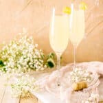 Two French 77 cocktails on a white marble board with a Champagne cork to the right and white flowers all around them.
