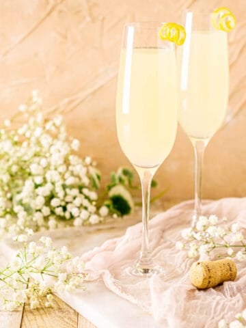 Two French 77 cocktails on a white marble board with a Champagne cork to the right and white flowers all around them.