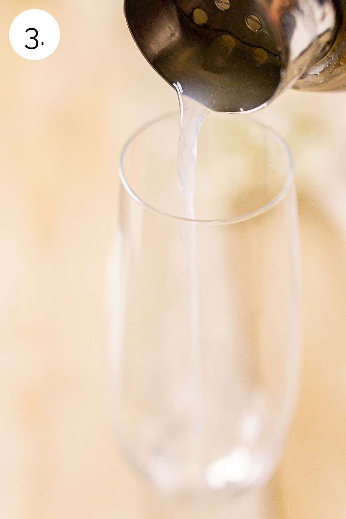 A cocktail shaker straining the lemon juice mixture into an empty Champagne flute on a wooden board.