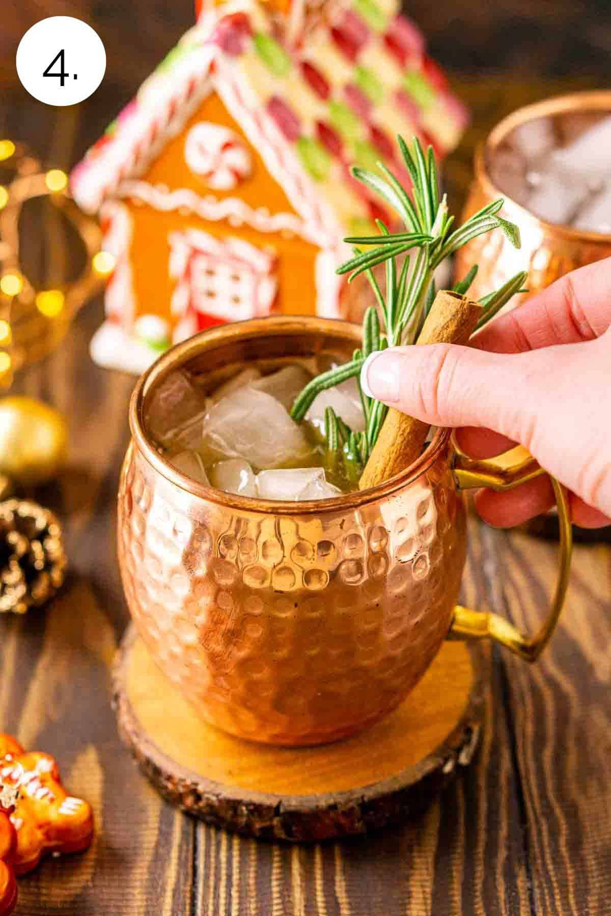 A hand adding a cinnamon stick and rosemary sprig to the copper mug as a garnish.