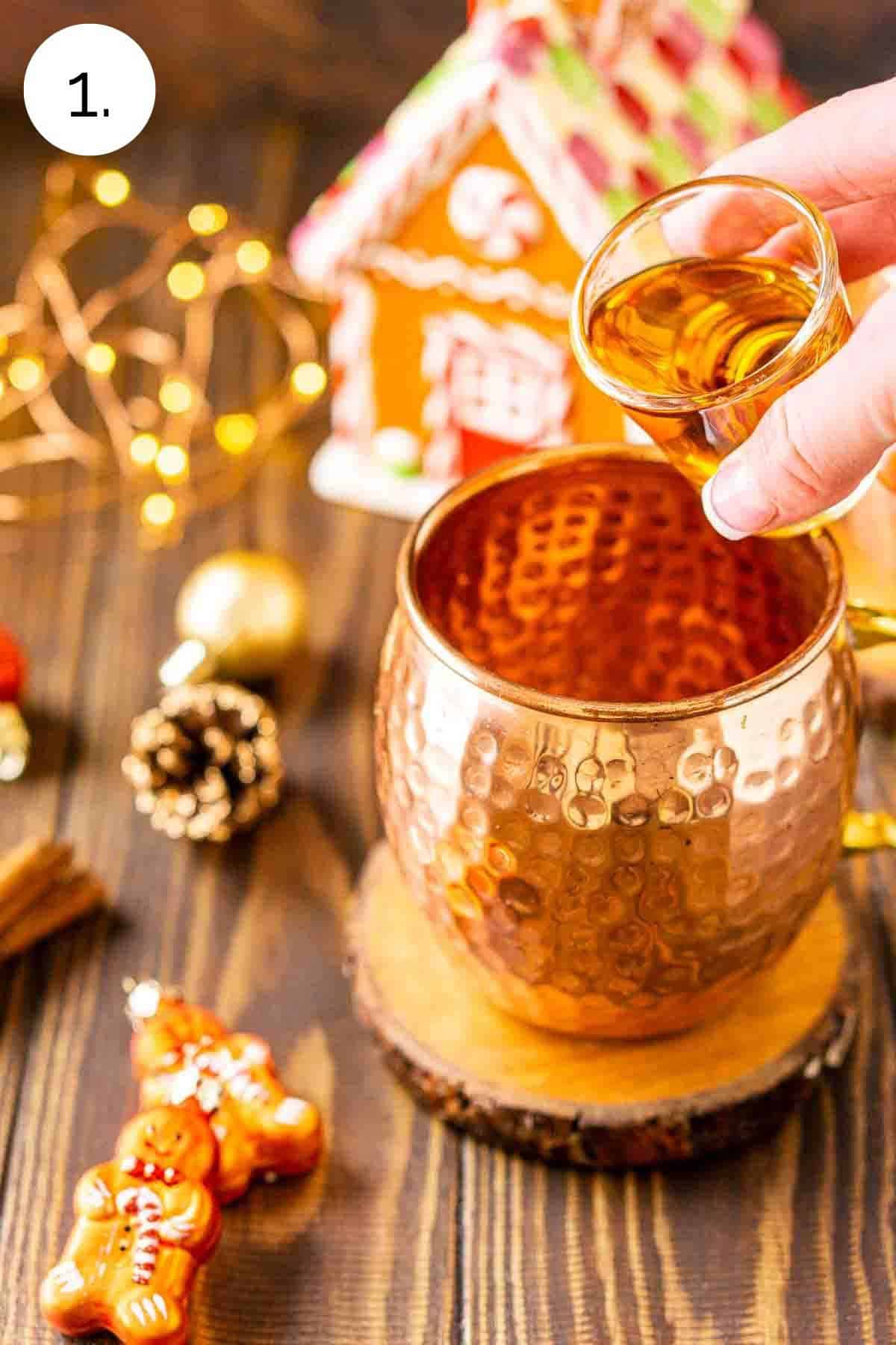 A hand pouring in a shot of bourbon into the copper mug with holiday decor all around.