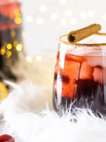 A wine glass filled with holiday sangria on a fluffy white piece of fabric with lights in the background and cranberries to the left.