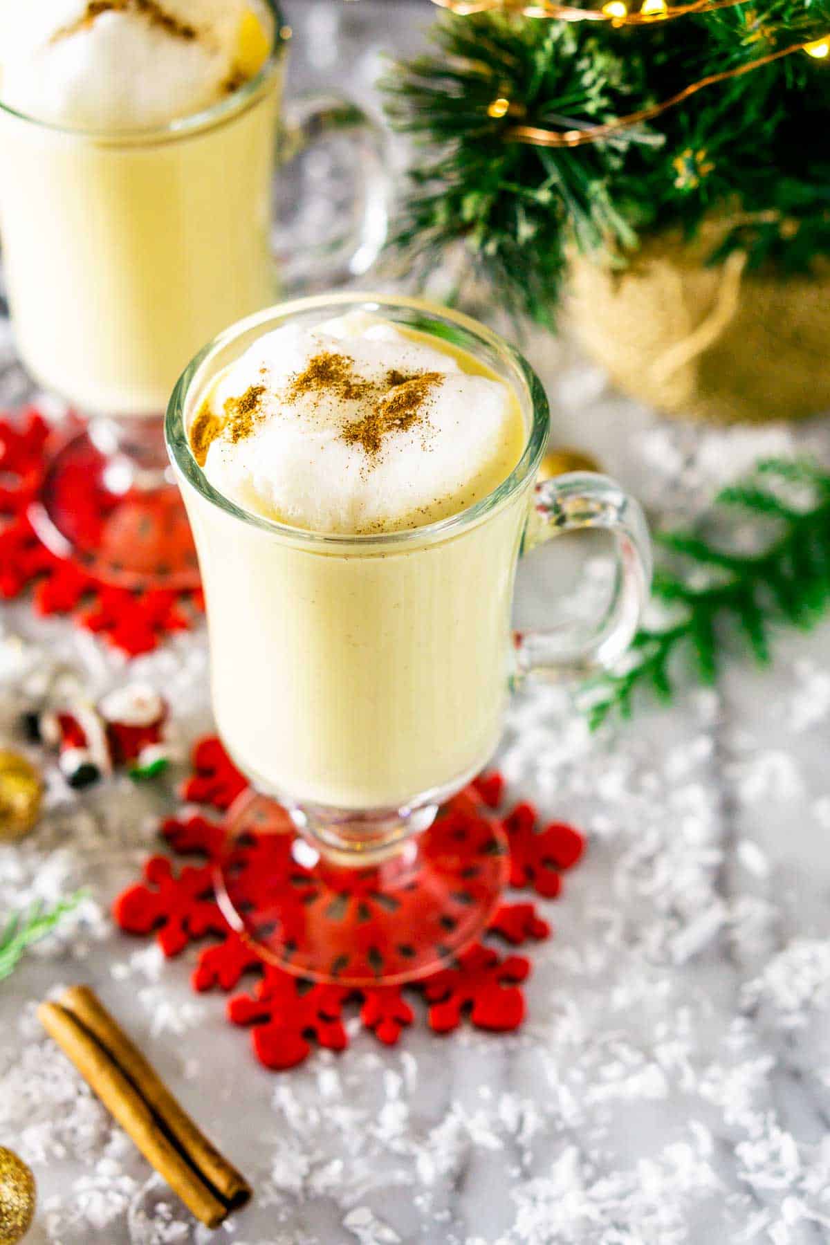 Two mugs of the maple eggnog on red coasters on top of a white countertop with fake snow and Christmas decor around them.