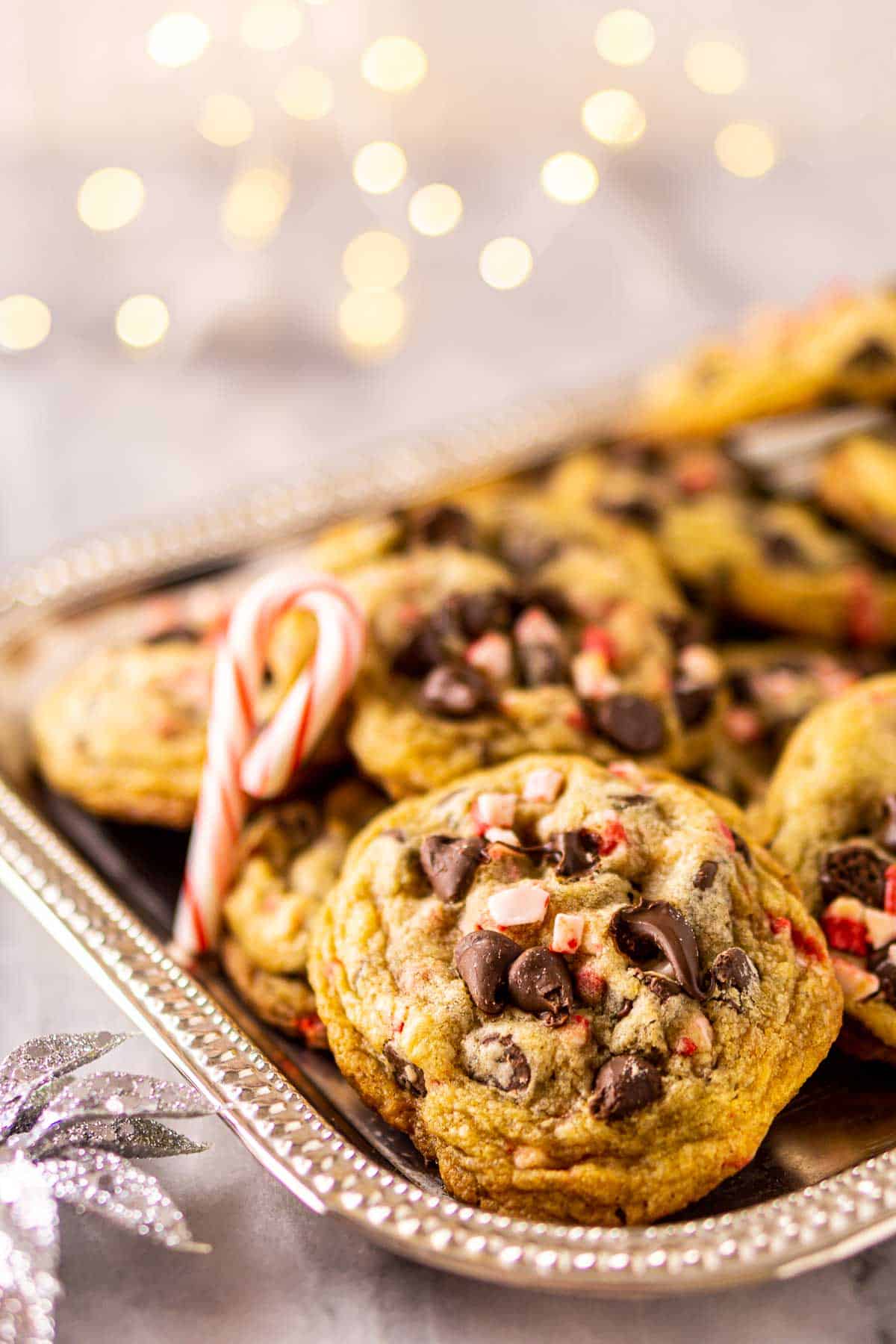 A close-up view of a peppermint chocolate chip cookie on a silver platter with white lights and a candy cane behind it.