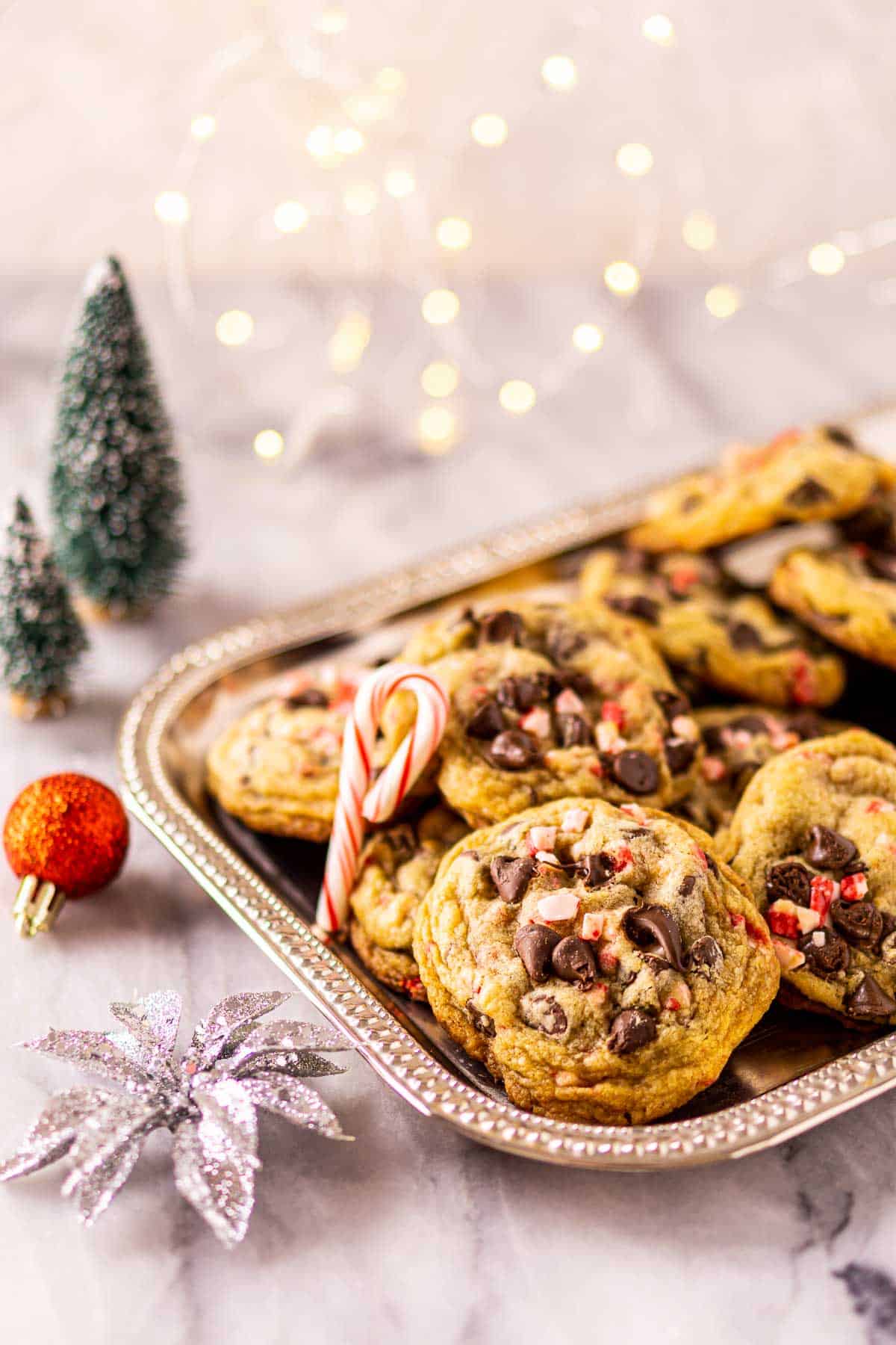 The peppermint chocolate chip cookies on a silver platter with sparkly lights in the background and holiday decor to the left.