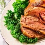 The smoked leg of lamb on green lettuce on top of a white platter after it's been carved into slices with rosemary and red wine in the background.