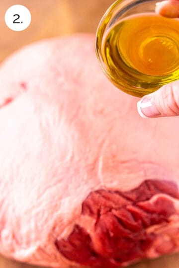 A hand holding a small bowl of olive oil to pour all over the leg of lamb roast on a brown cutting board.