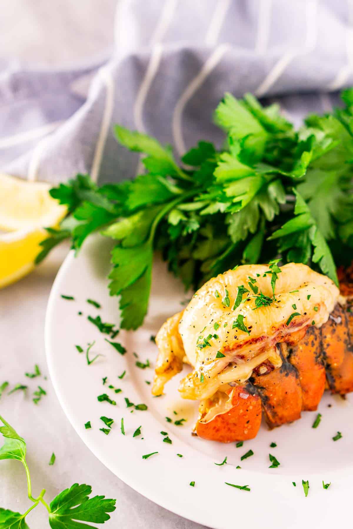 A smoked lobster tail on a white plate with a bunch of parsley to the side and a blue and white napkin behind it.