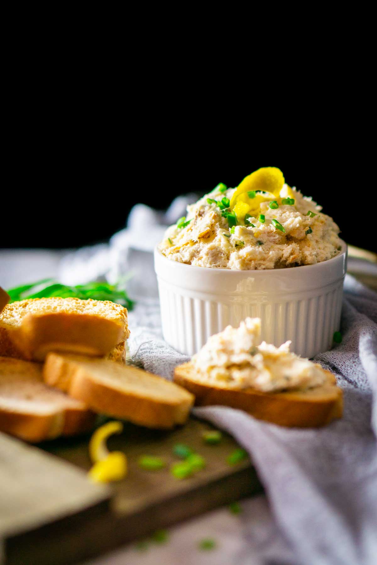 A white ramekin filled with the smoked trout dip against a black background on a wooden tray with lemon peel, chives and crostini.