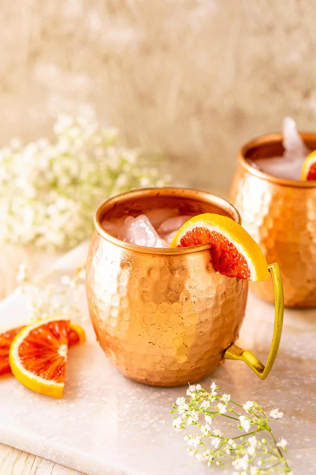 A close-up view from the side of the blood orange Moscow mule on a white marble tray with a small bundle of flowers and blood orange slices in front.