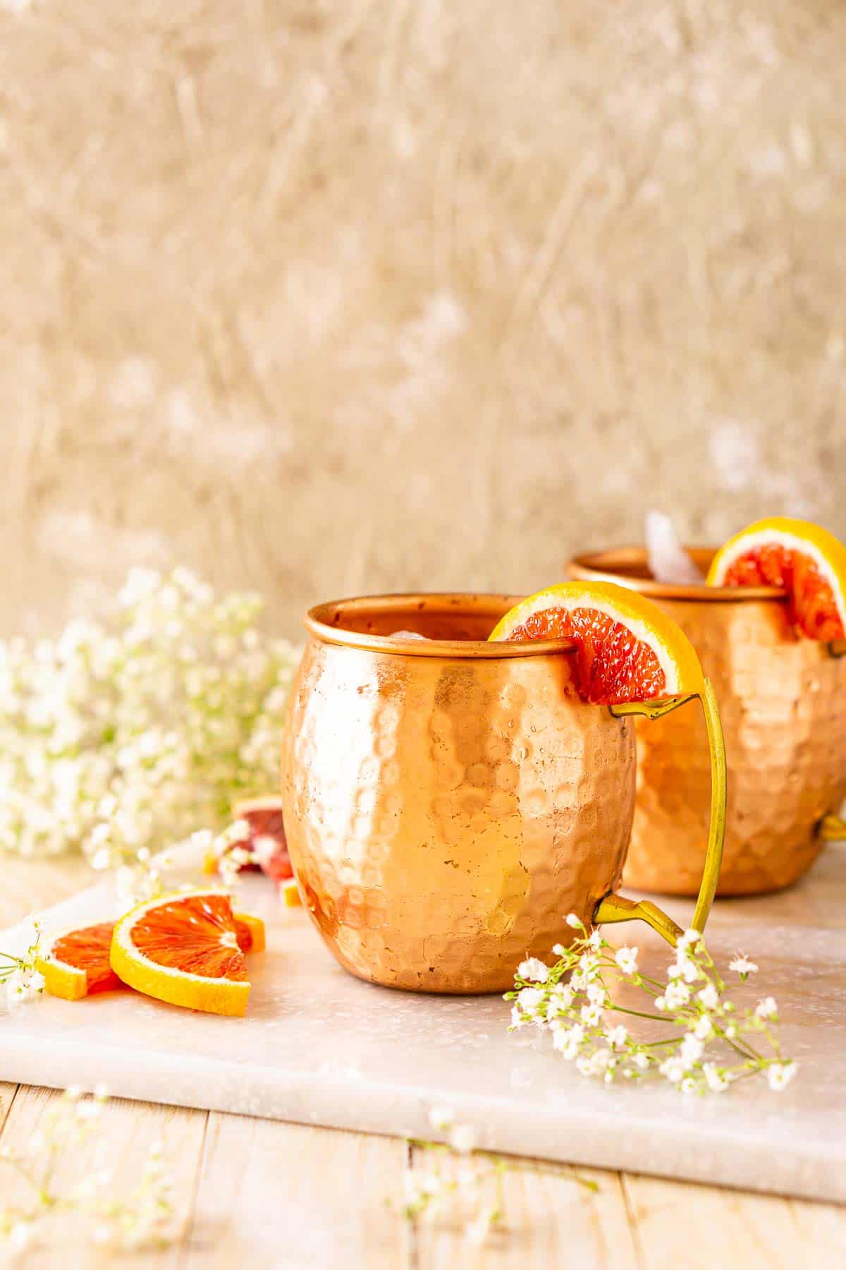 Looking straight on to two blood orange Moscow mules on a white marble tray with blood orange slices and flowers surrounding them.