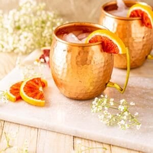 Looking down on a blood orange Moscow mule on a white marble tray with slices of blood orange and white flowers to the side.