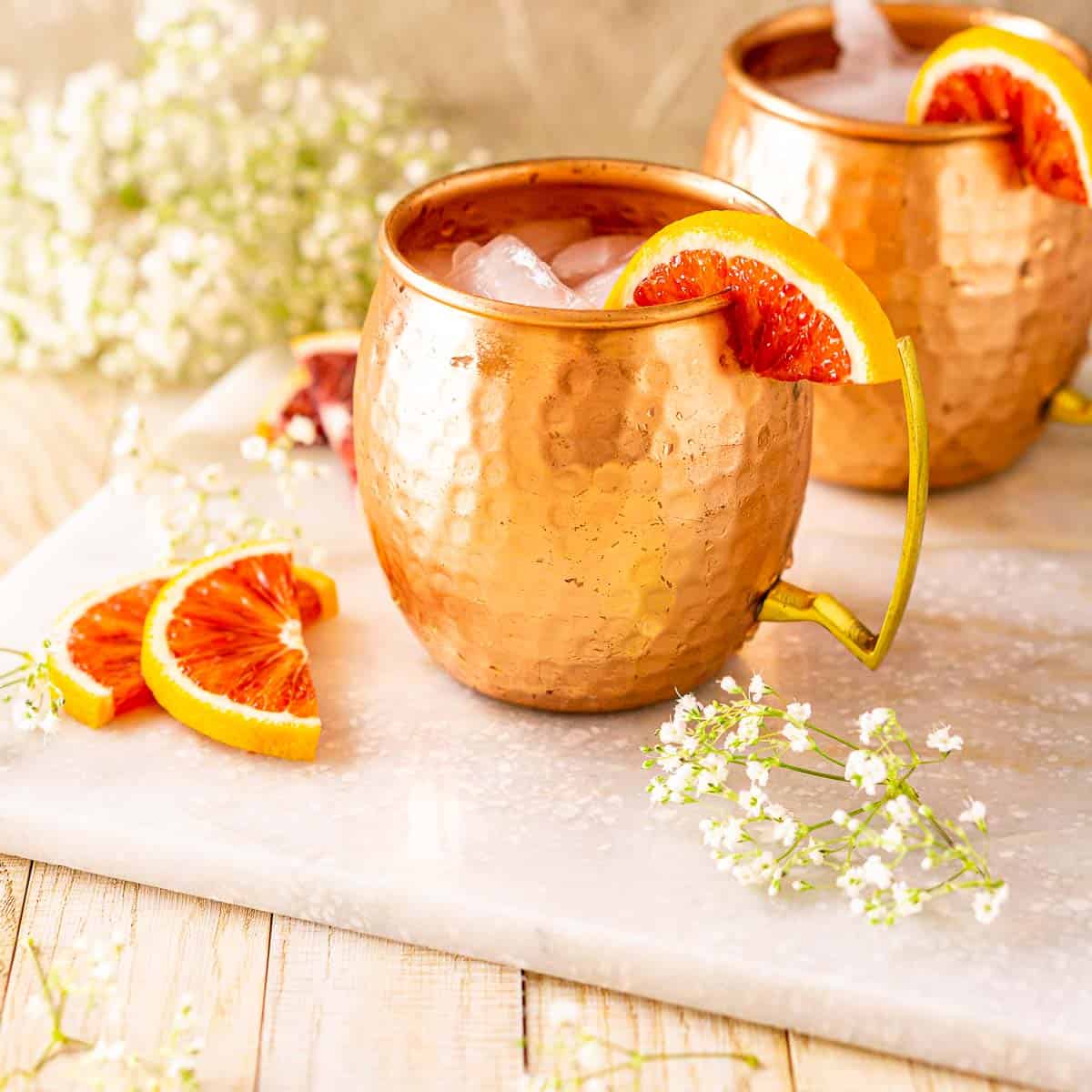 Two blood orange mule cocktails on a white marble tray with flowers around them.