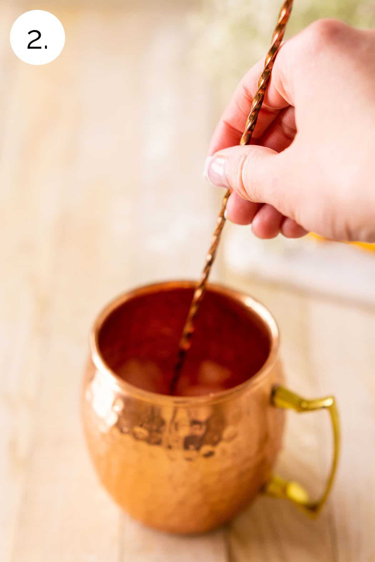 A hand stirring the drink in a copper mug with a long bar spoon on a cream-colored wooden surface.