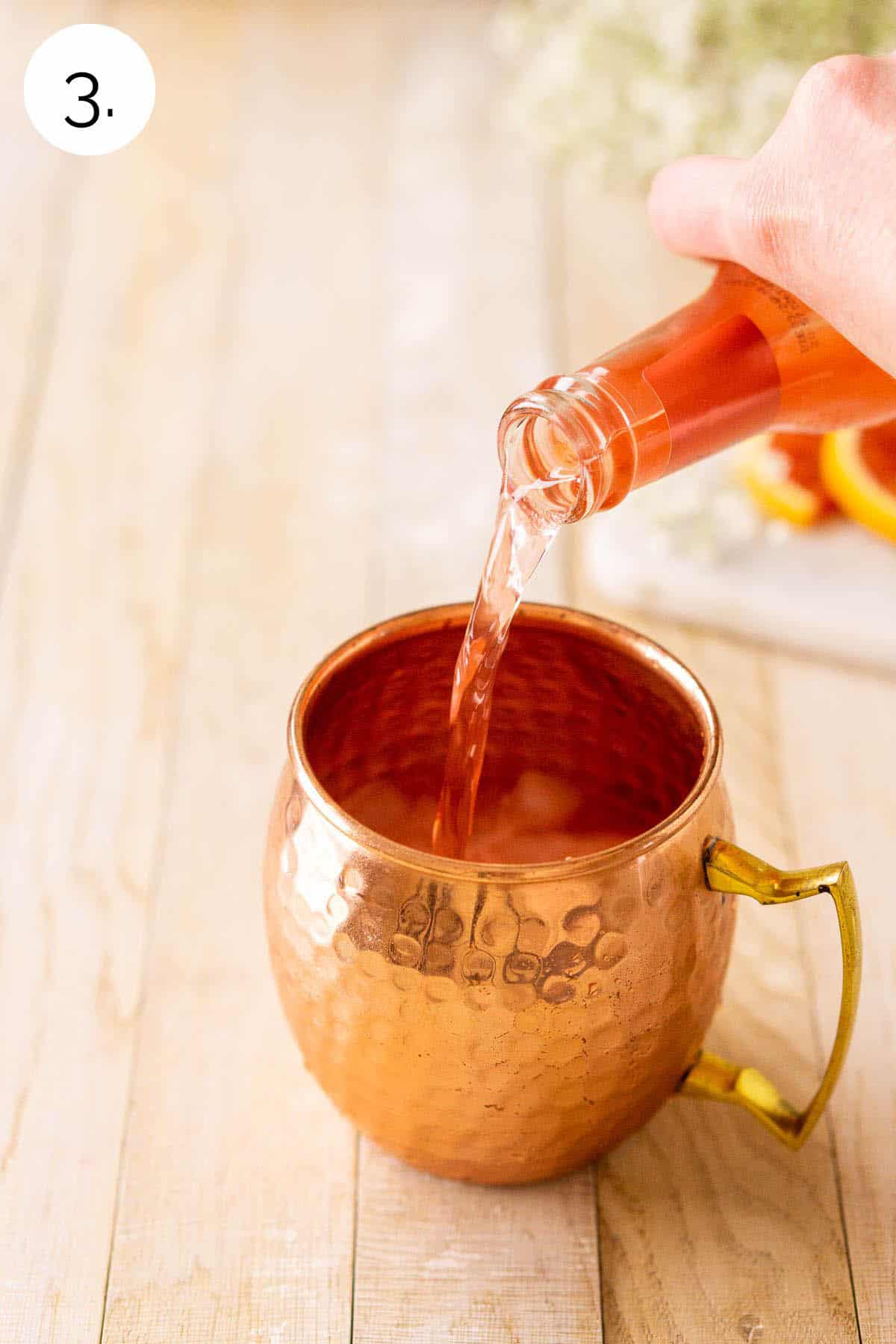 A hand pouring a bottle of blood orange ginger beer into the copper mug on a cream-colored wooden surface.