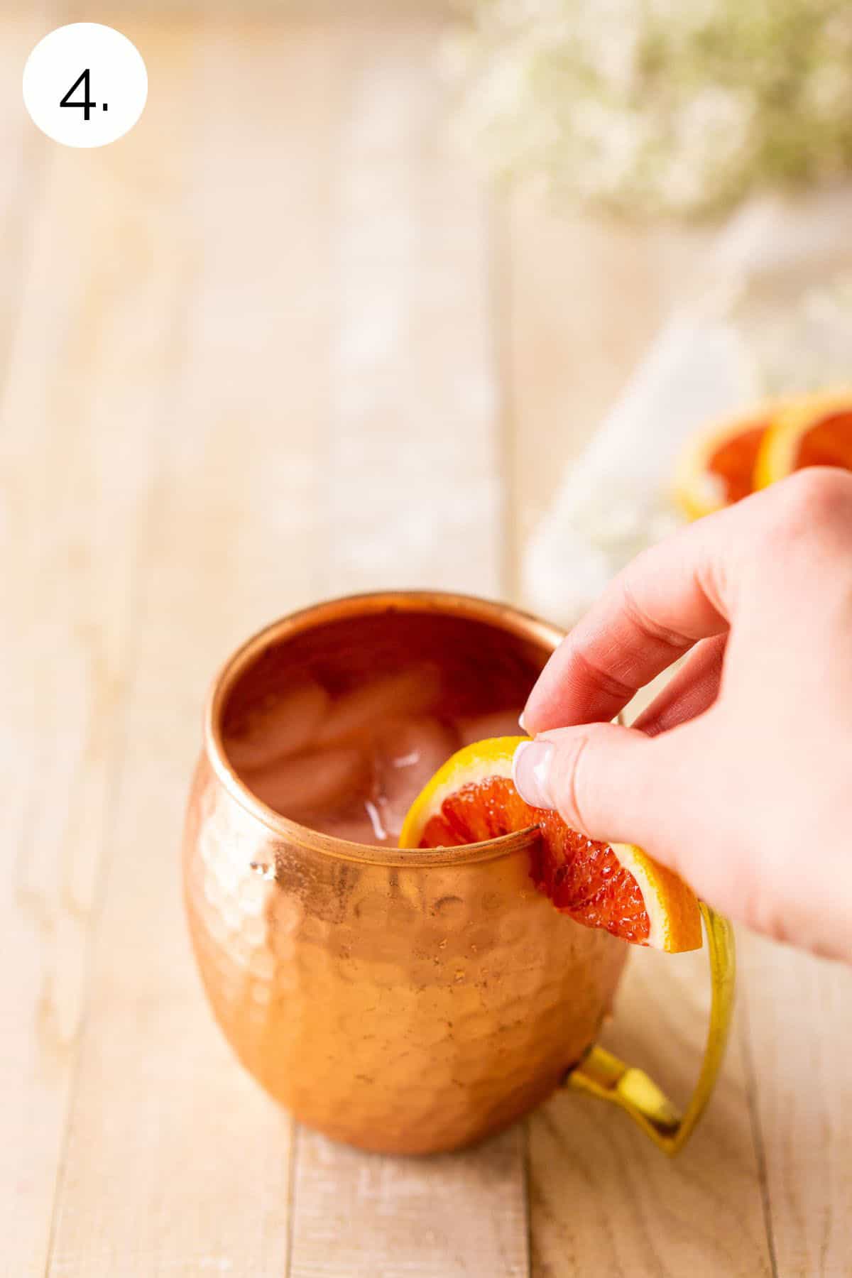 A hand placing a blood range slice on the rim of the copper mug on a cream-colored wooden surface.