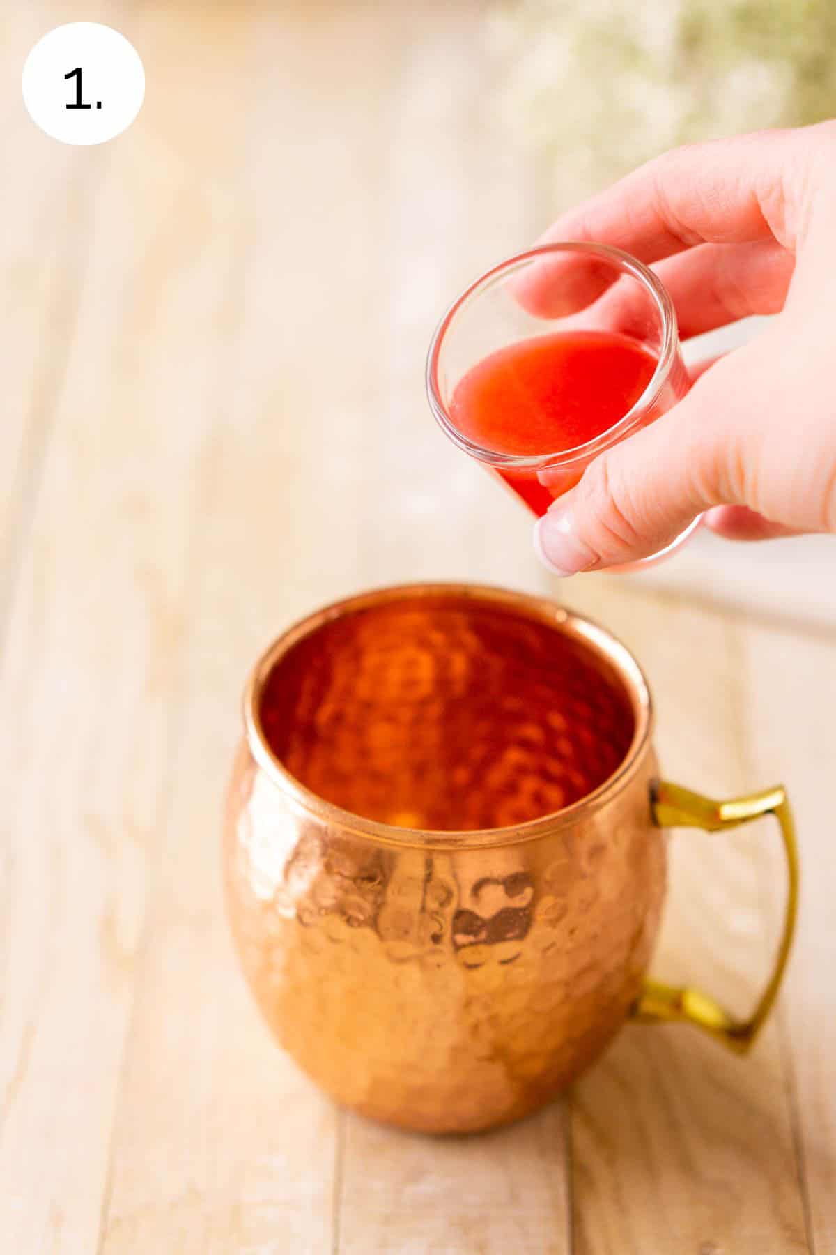A hand pouring a shot glass full of the blood orange juice into the copper mug on a cream-colored wooden surface.