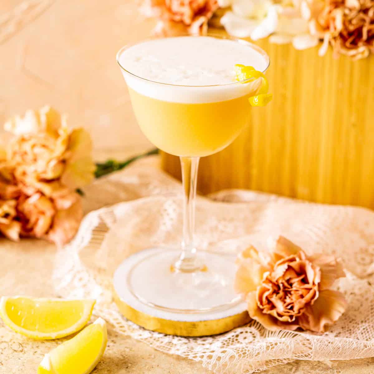 A Boston sour on a white and gold coaster with lemon wedges and flowers around it.