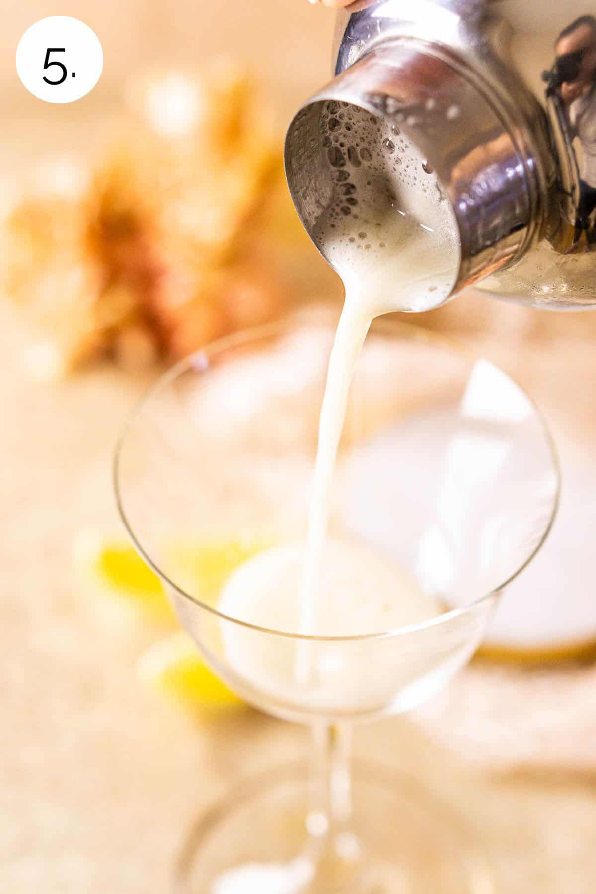 Straining the drink from the cocktail shaker into a sour glass on a cream-colored marble surface.