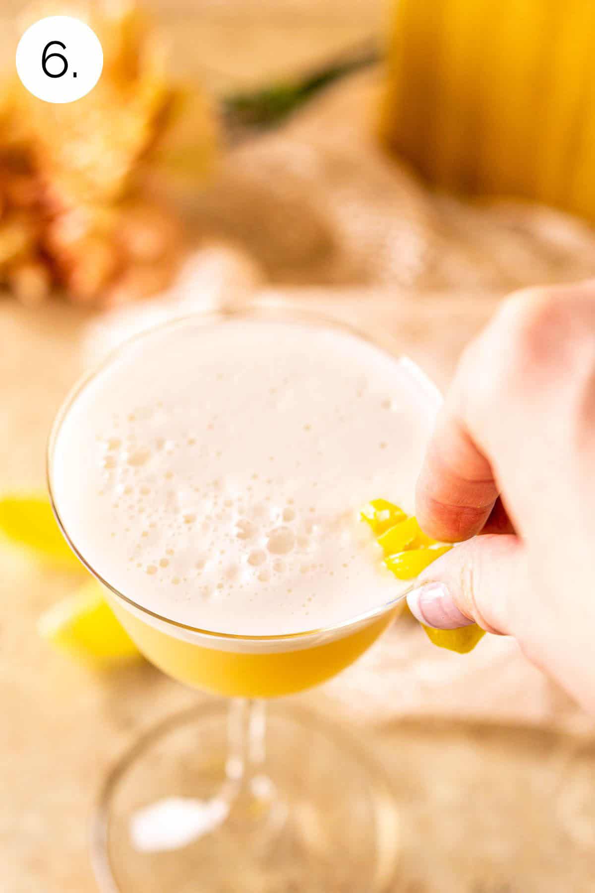 A hand placing a lemon twist on the side of the serving glass after straining the drink.