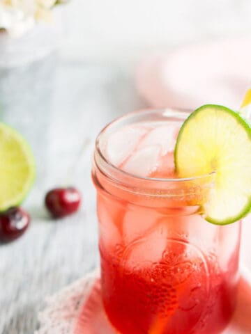 A bourbon cherry limemade on a pink napkin with white flowers, fresh cherries and a lime in the background.