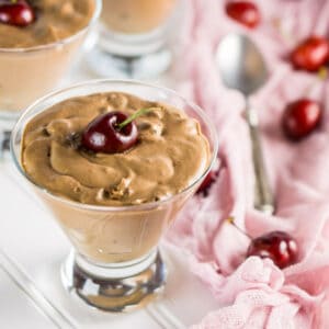 A dessert glass filled with the chocolate cherry cream on a white surface with a pink decorative clothe next to it.