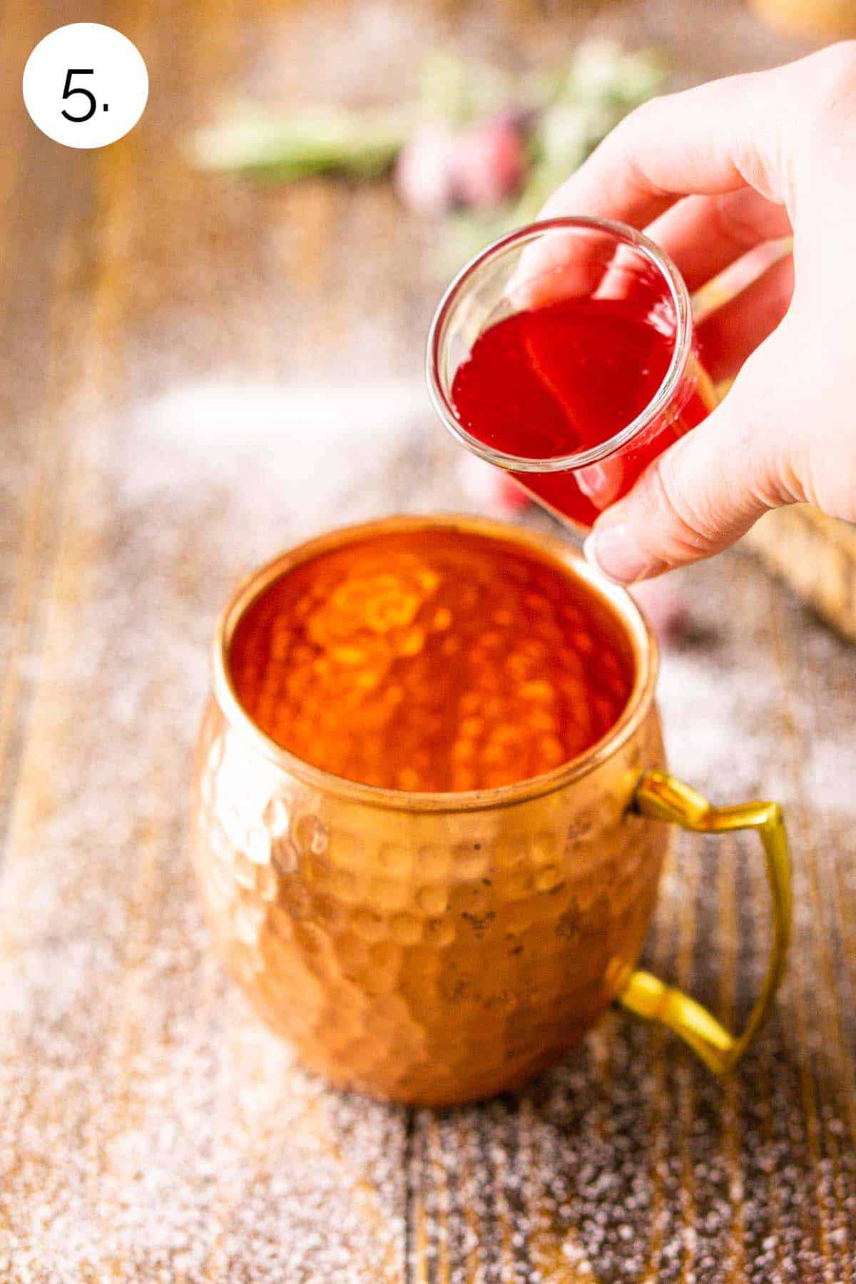 A hand pouring a shot glass of the cranberry syrup into a copper mug on a brown wooden surface.