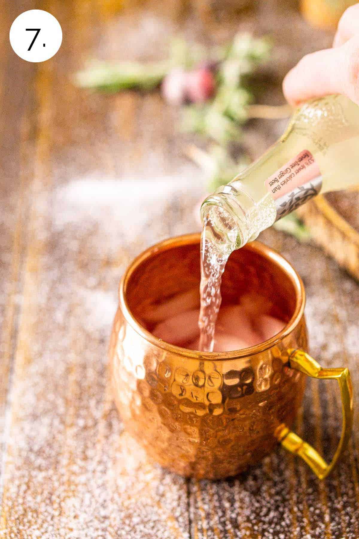 A hand pouring a bottle of ginger beer into the copper mug on a brown wooden surface.
