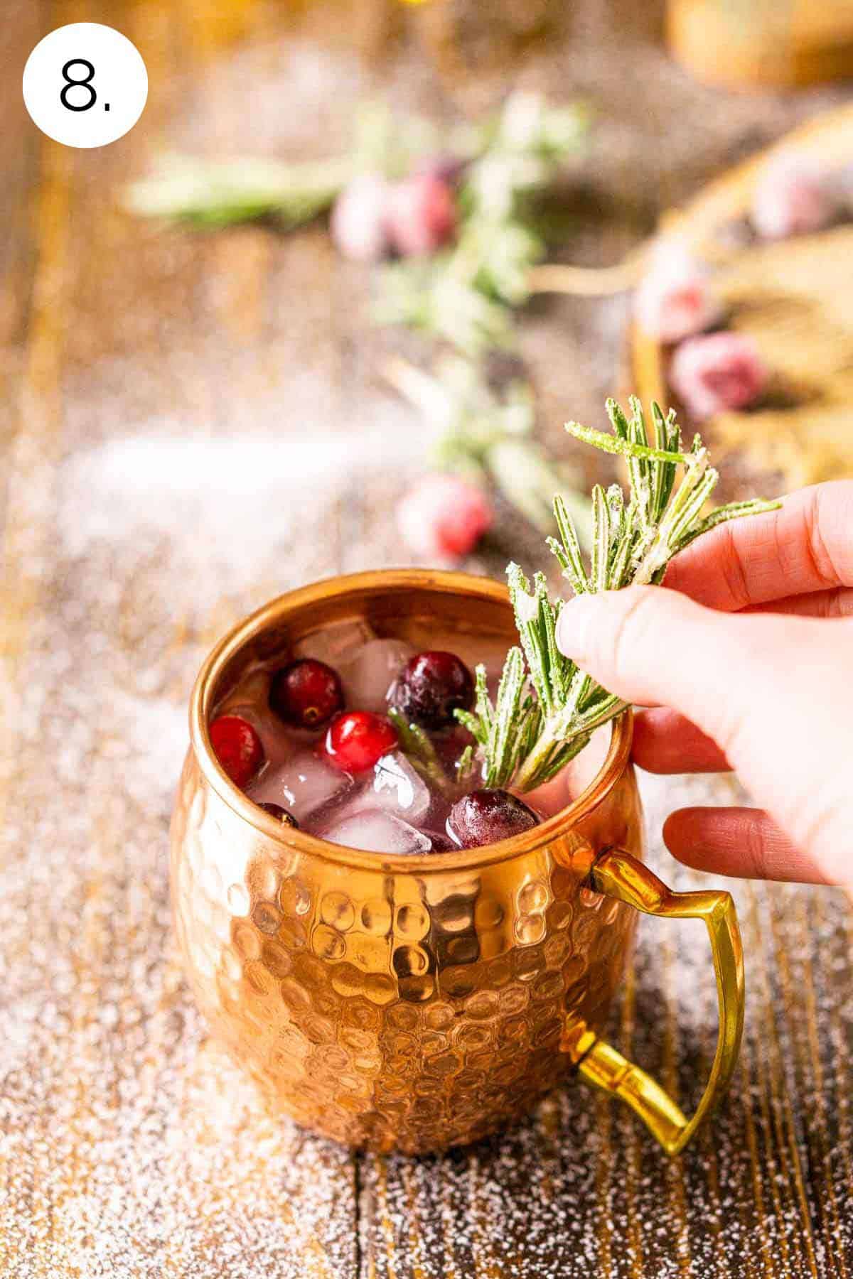 A hand placing a sugared rosemary sprig into the copper mug on a brown wooden surface.
