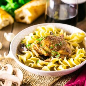 Coq au vin in a white bowl on burlap with mushrooms to the side and a loaf of crusty bread in the background.