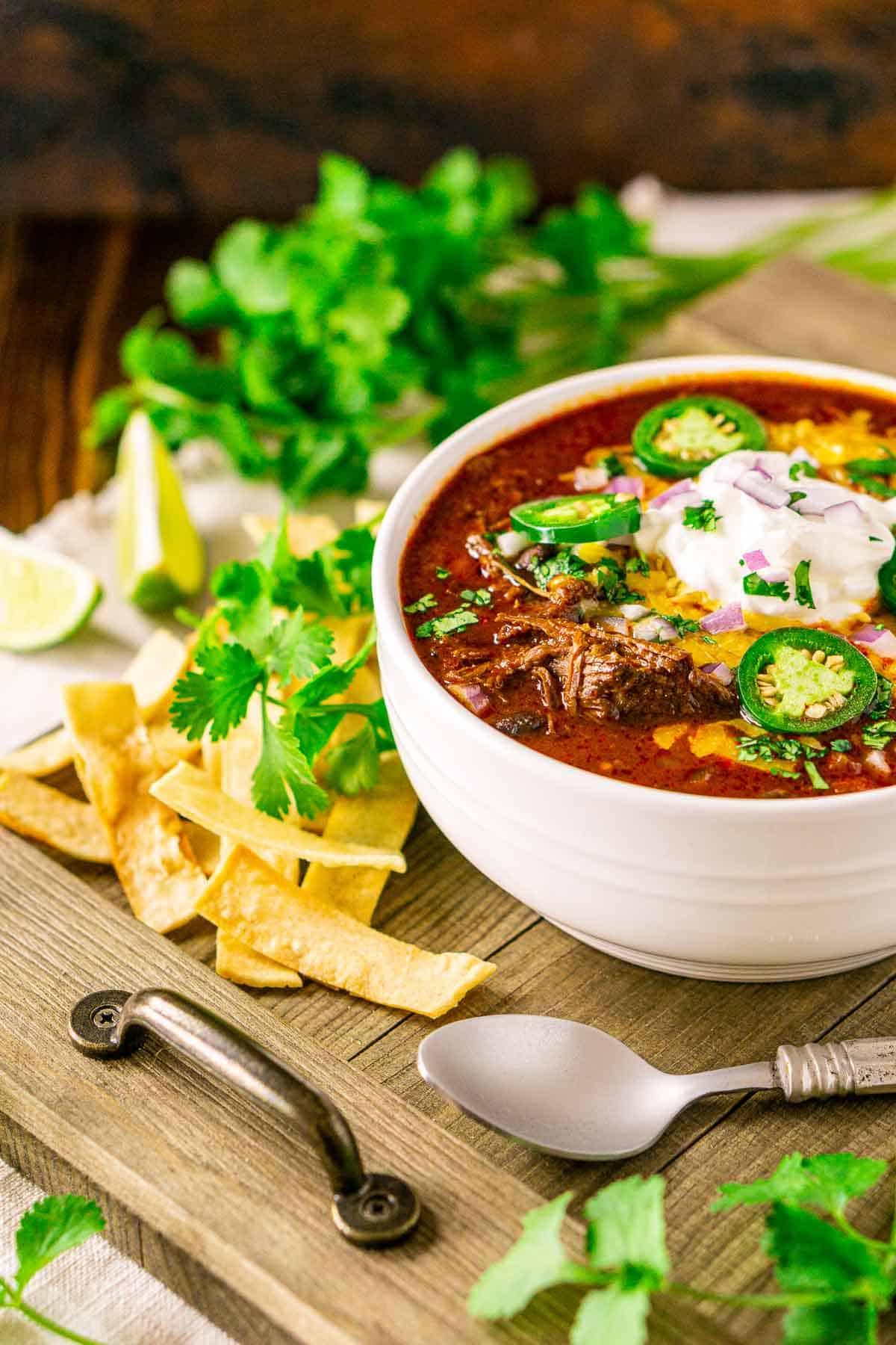 A bowl of the Dutch oven chili on a wooden serving tray with cilantro, tortilla strips and lime slices in the background.