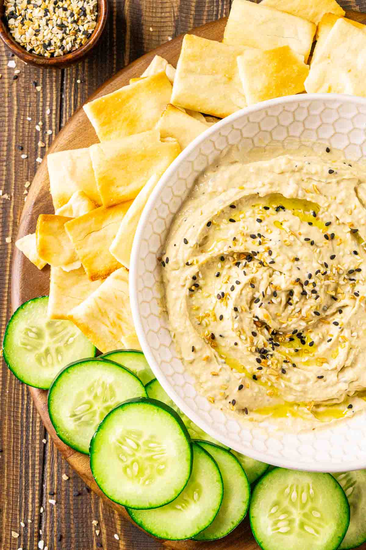 An aerial view of a bowl of everything bagel hummus in a cream-colored bowl on a wooden surface with cucumbers and pita chips.