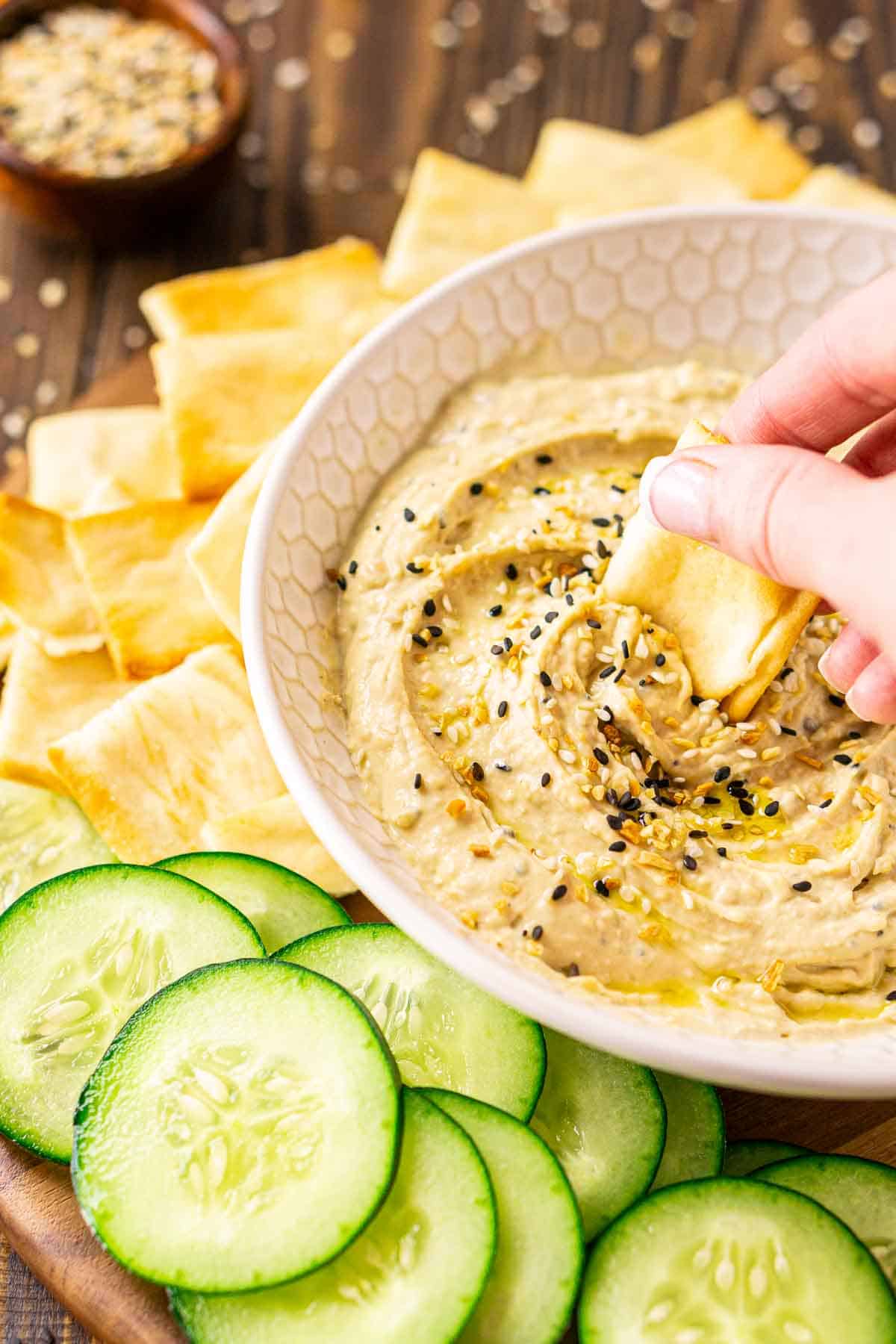 A hand dipping a pita chip into the bowl of everything bagel hummus with cucumber slices in front.