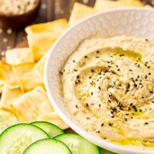 A cream-colored bowl with everything bagel hummus on a wooden platter with cucumber slices and pita chips scattered around it.