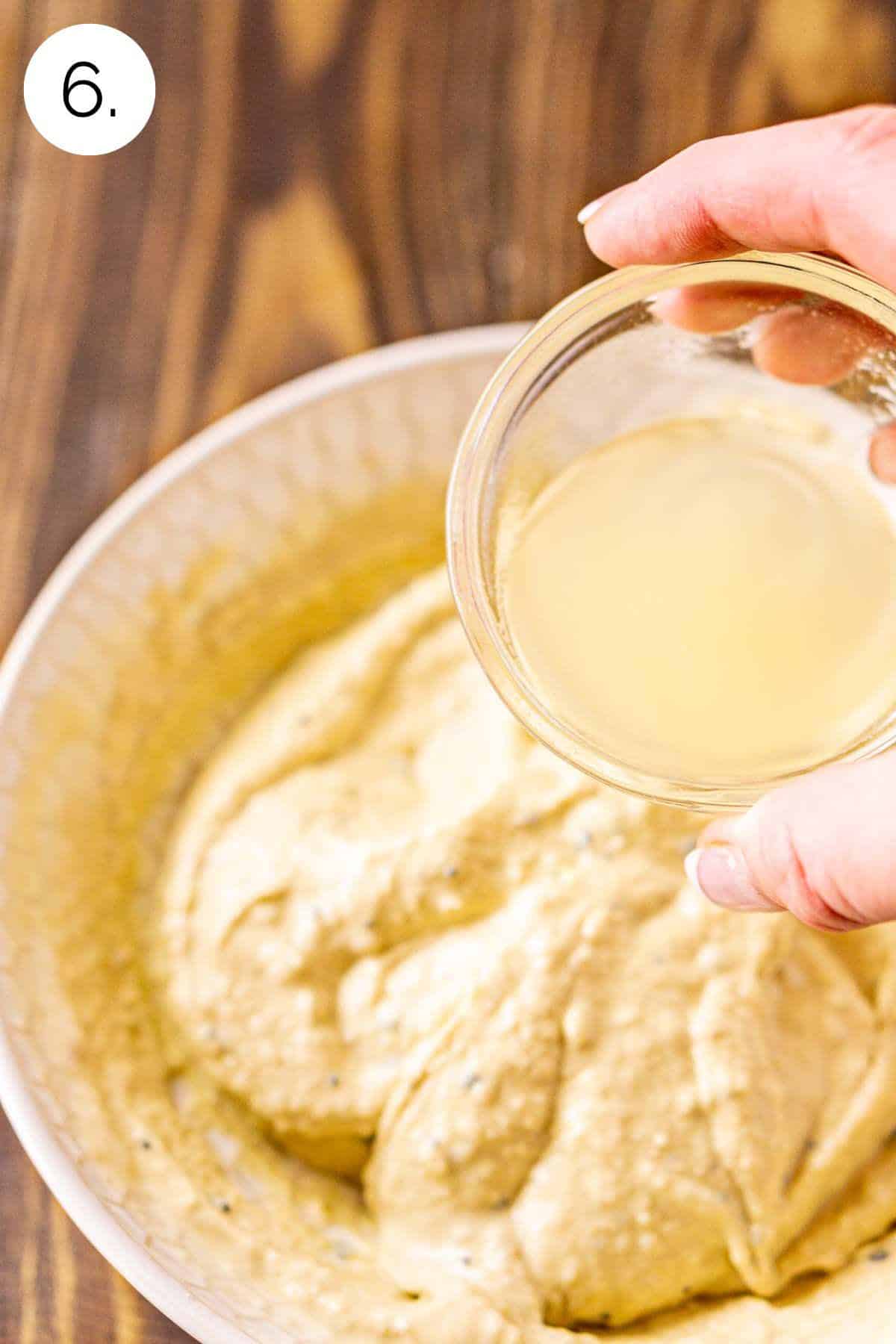 A hand holding a small bowl of the reserved chickpea liquid as it's about to pour it into the hummus to thin out.