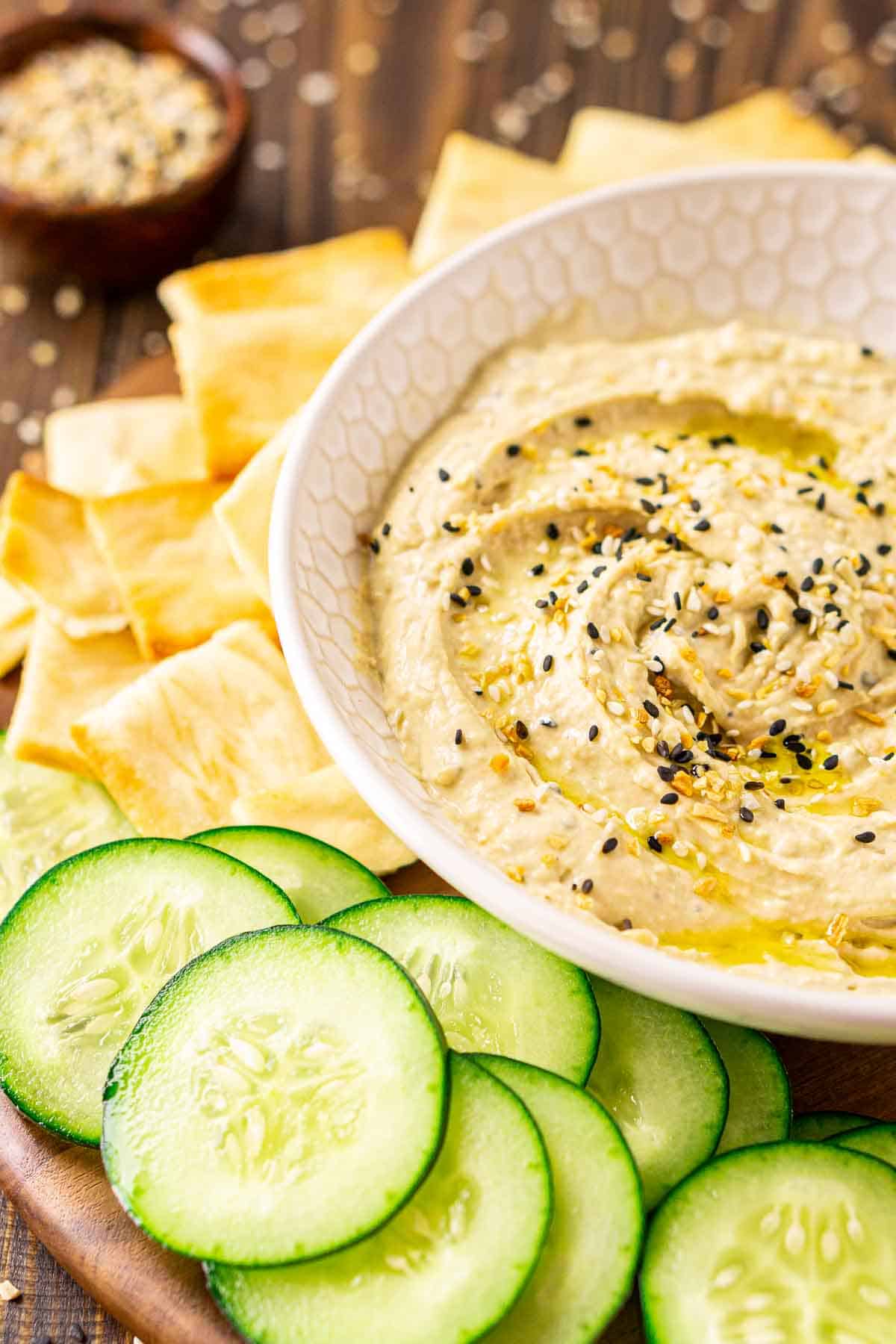 A cream-colored bowl with everything bagel hummus on a wooden platter with cucumber slices and pita chips scattered around it.