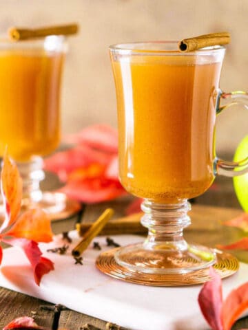 Two glasses of the homemade apple cider on a marble tray with spices and fall leaves around them.