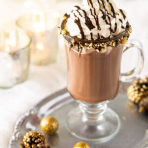 A glass mug of the homemade hot cocoa on a silver platter with sparkling holiday decor around it.