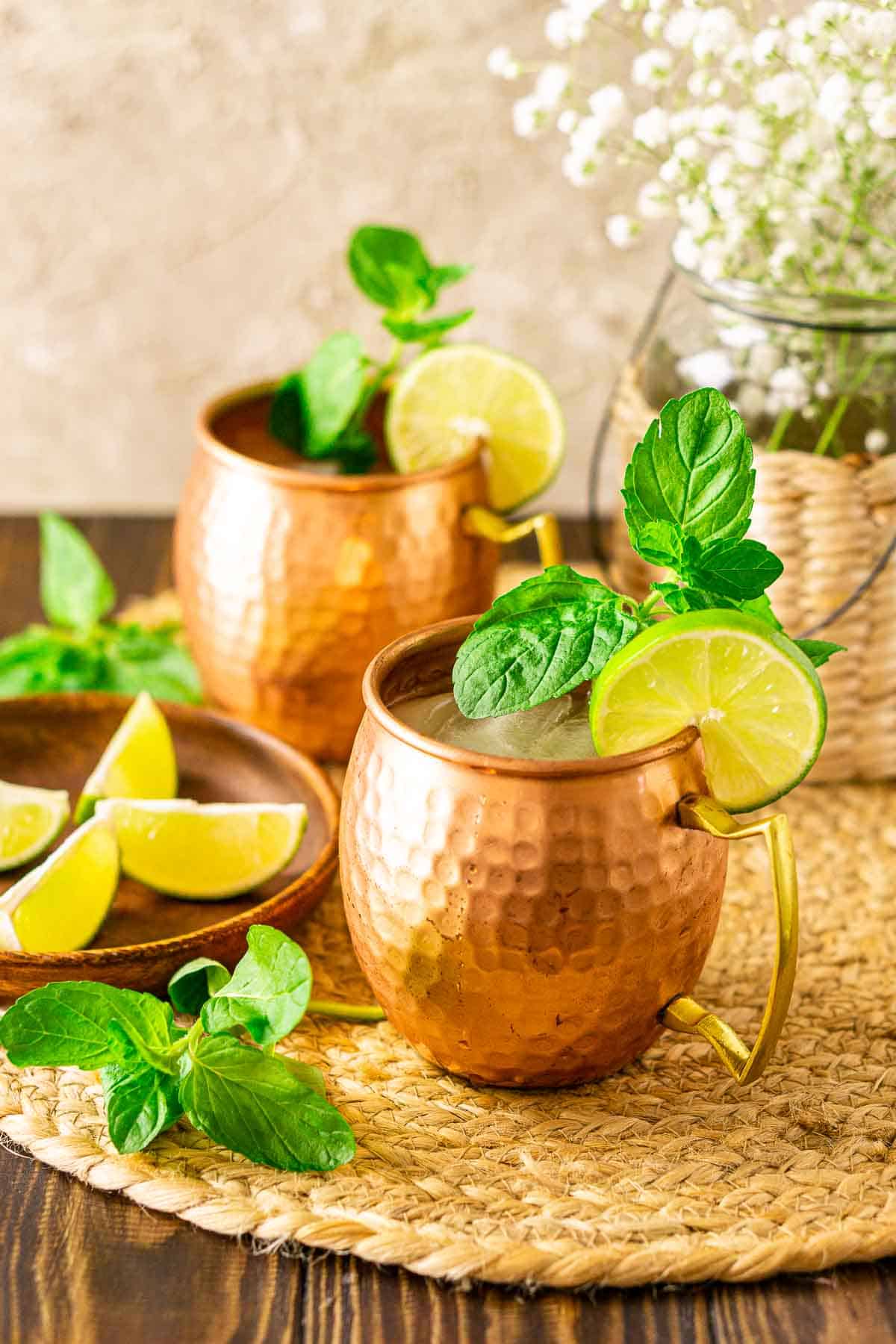 Looking down on two Kentucky mule cocktails on a straw placement with a wooden plate filled with limes on the left.